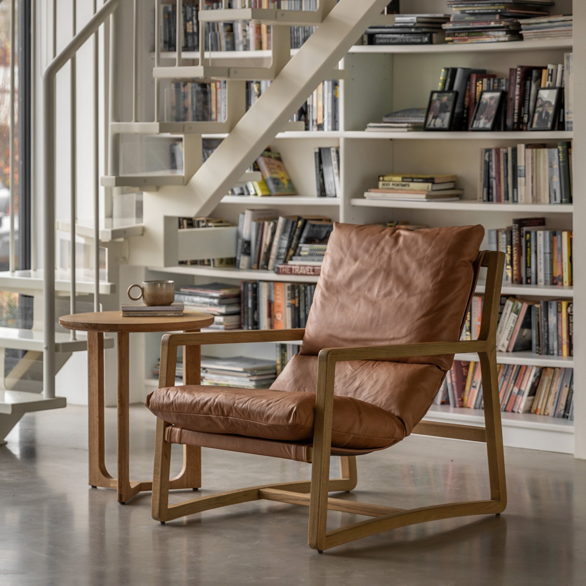 Tan leather and wooden armchair in a modern living room with bookshelves and a staircase.