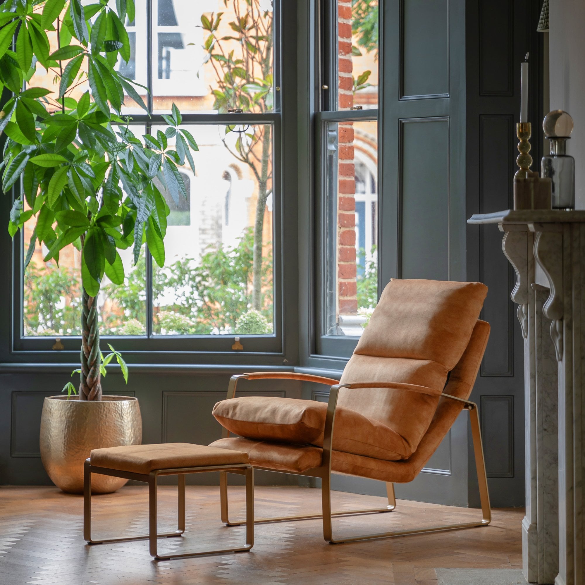 A luxurious tan lounge chair with an ottoman in a well-lit living room by a large window.