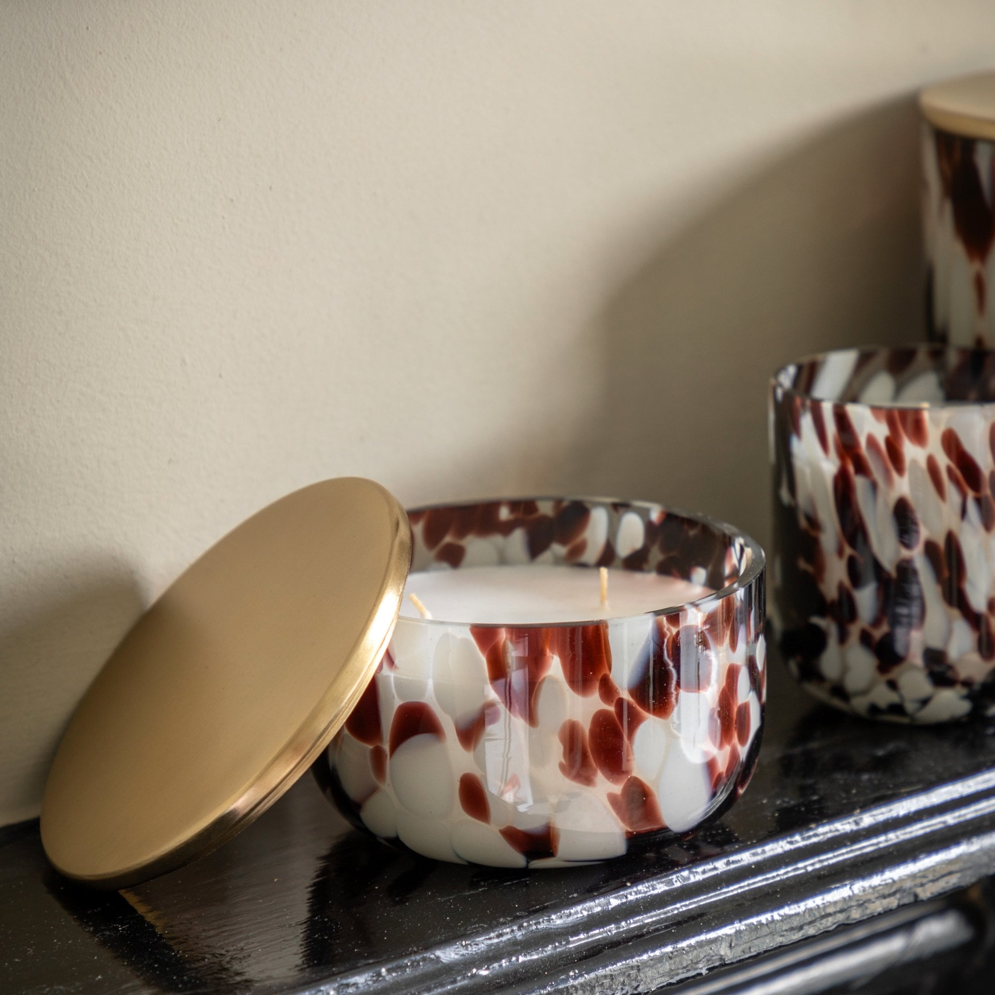A short, wide glass candle in white with deep red and black splotches, lid ajar.