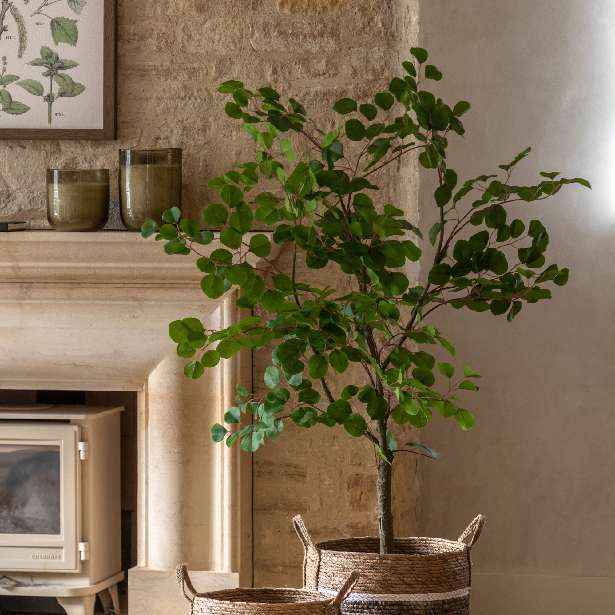 A faux eucalyptus tree in a woven basket, positioned next to a rustic fireplace.