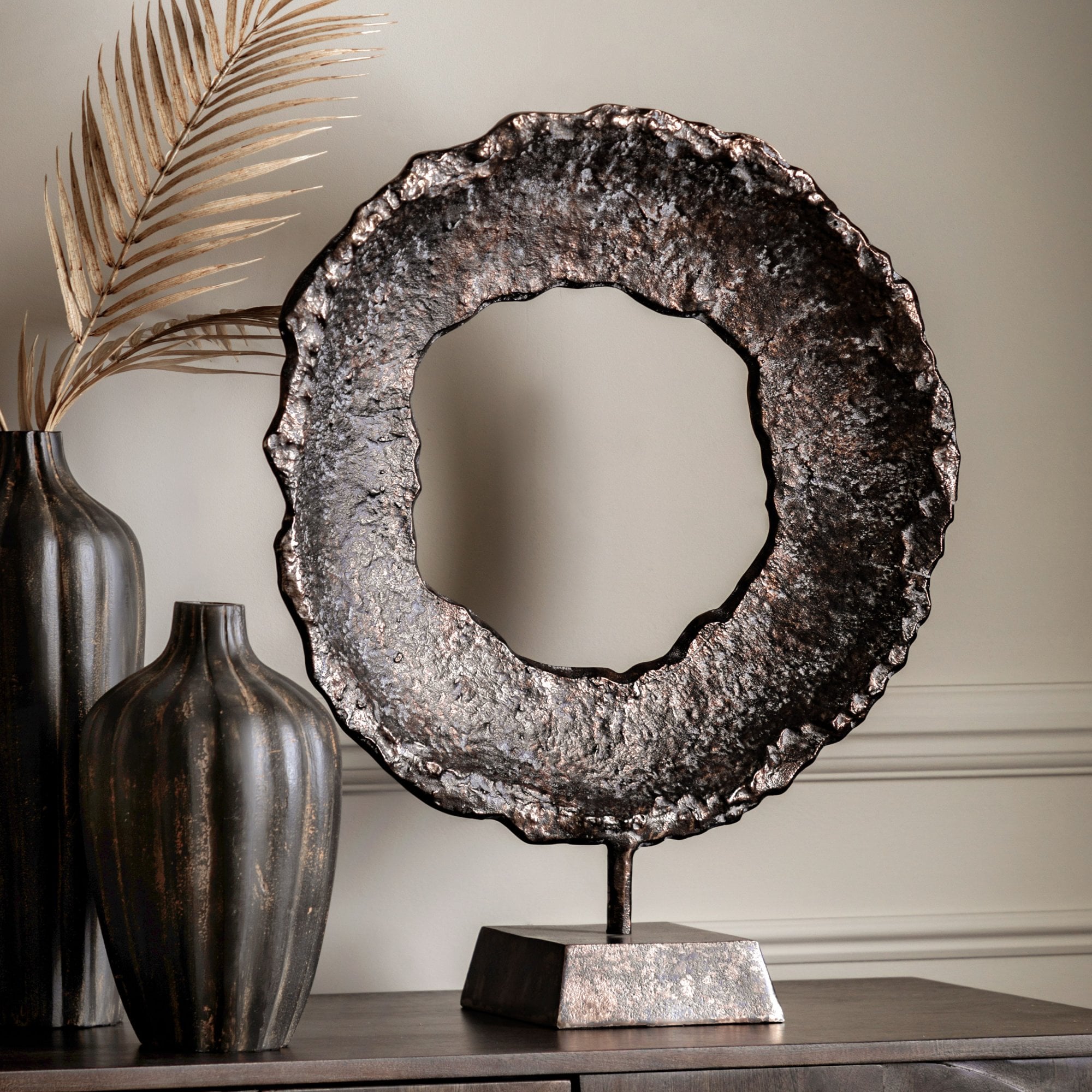 A rustic, bronze-colored metal ring sculpture on a stand, flanked by two dark vases and a dried palm leaf.