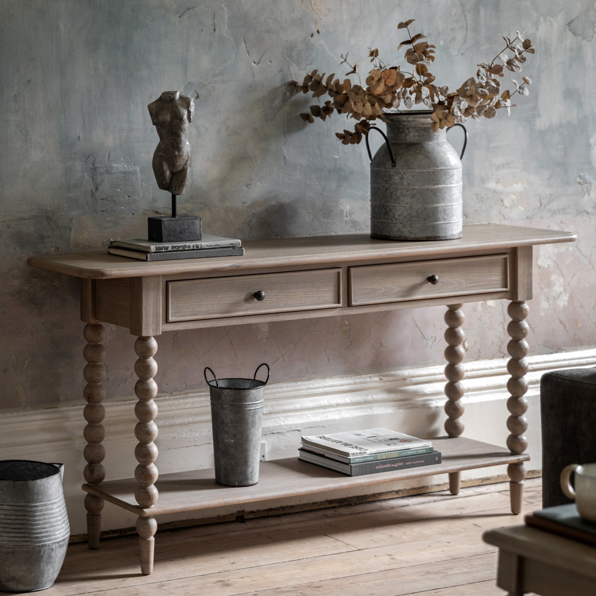 Light wood console table with two drawers, a bottom shelf, and bobbin legs, styled in a living room.