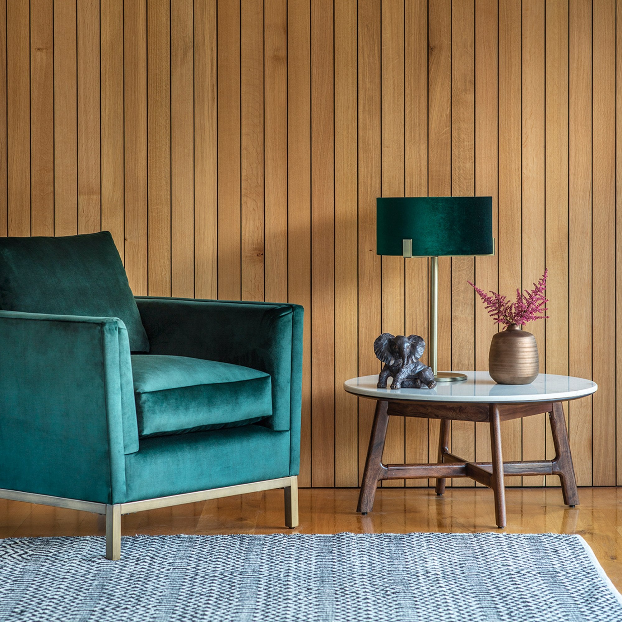 A green velvet armchair next to a round coffee table with decor against a wood-paneled wall.