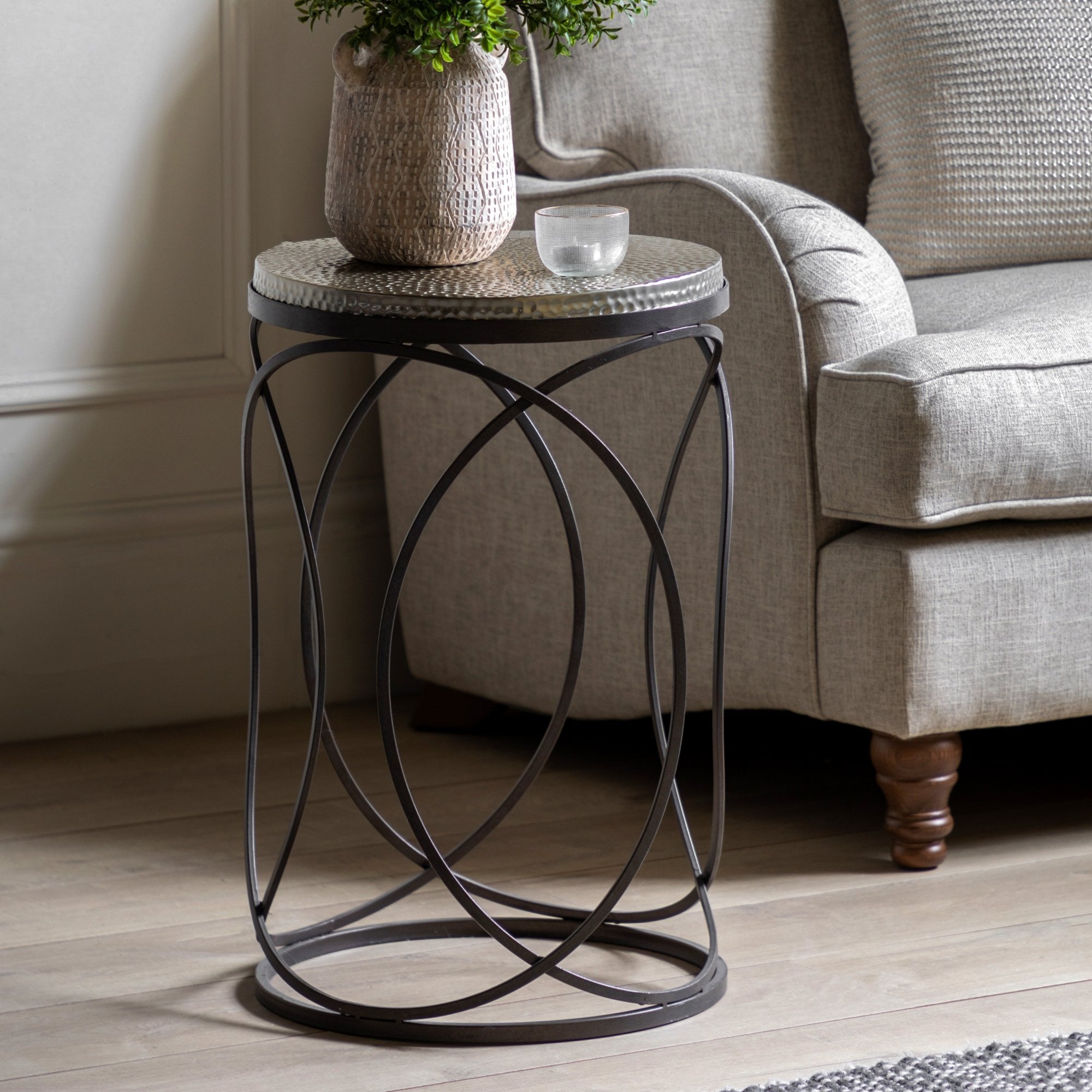 Round side table with a hammered metal top and black metal geometric base next to a gray sofa.