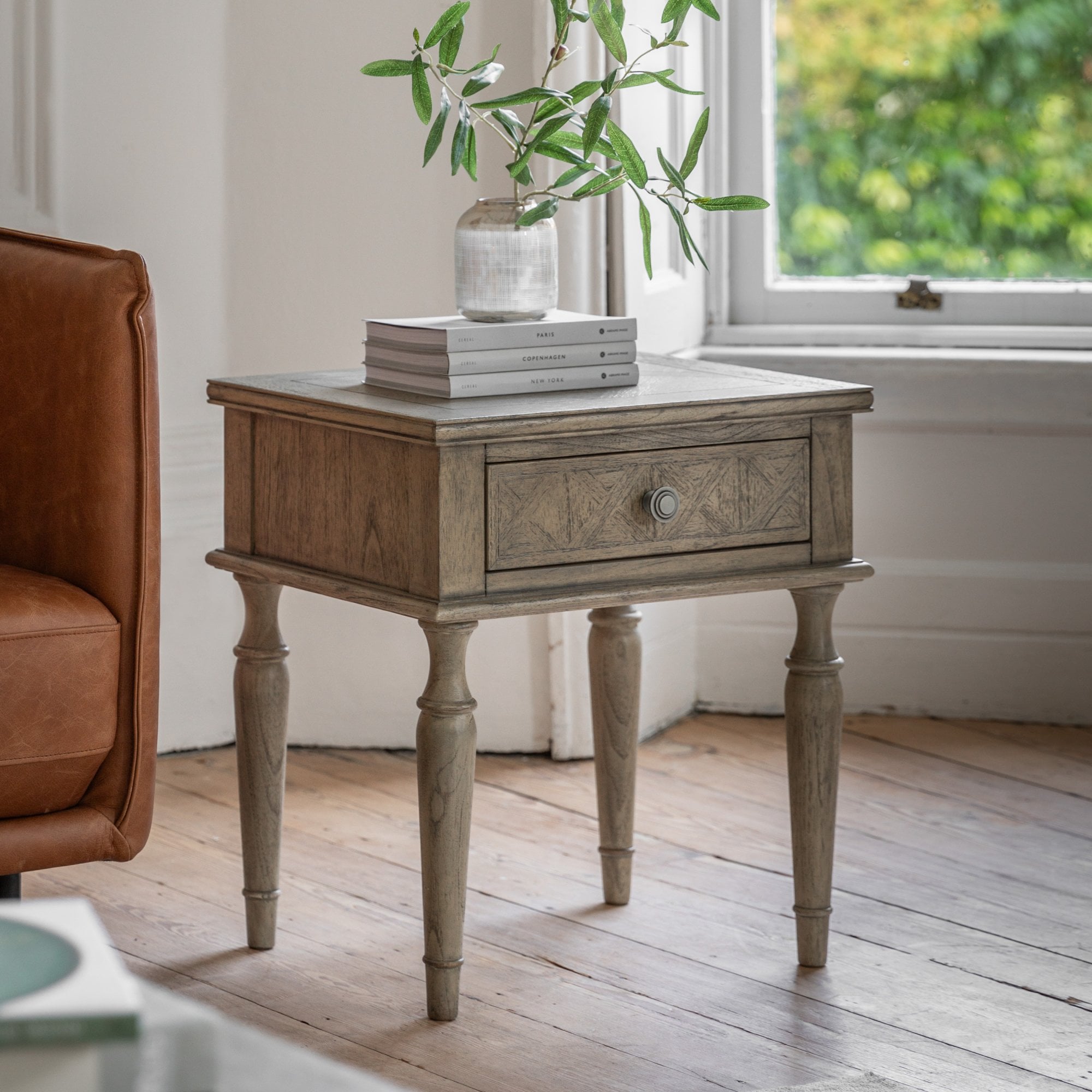 Distressed grey bedside table with a single drawer, and four turned legs, next to a window.
