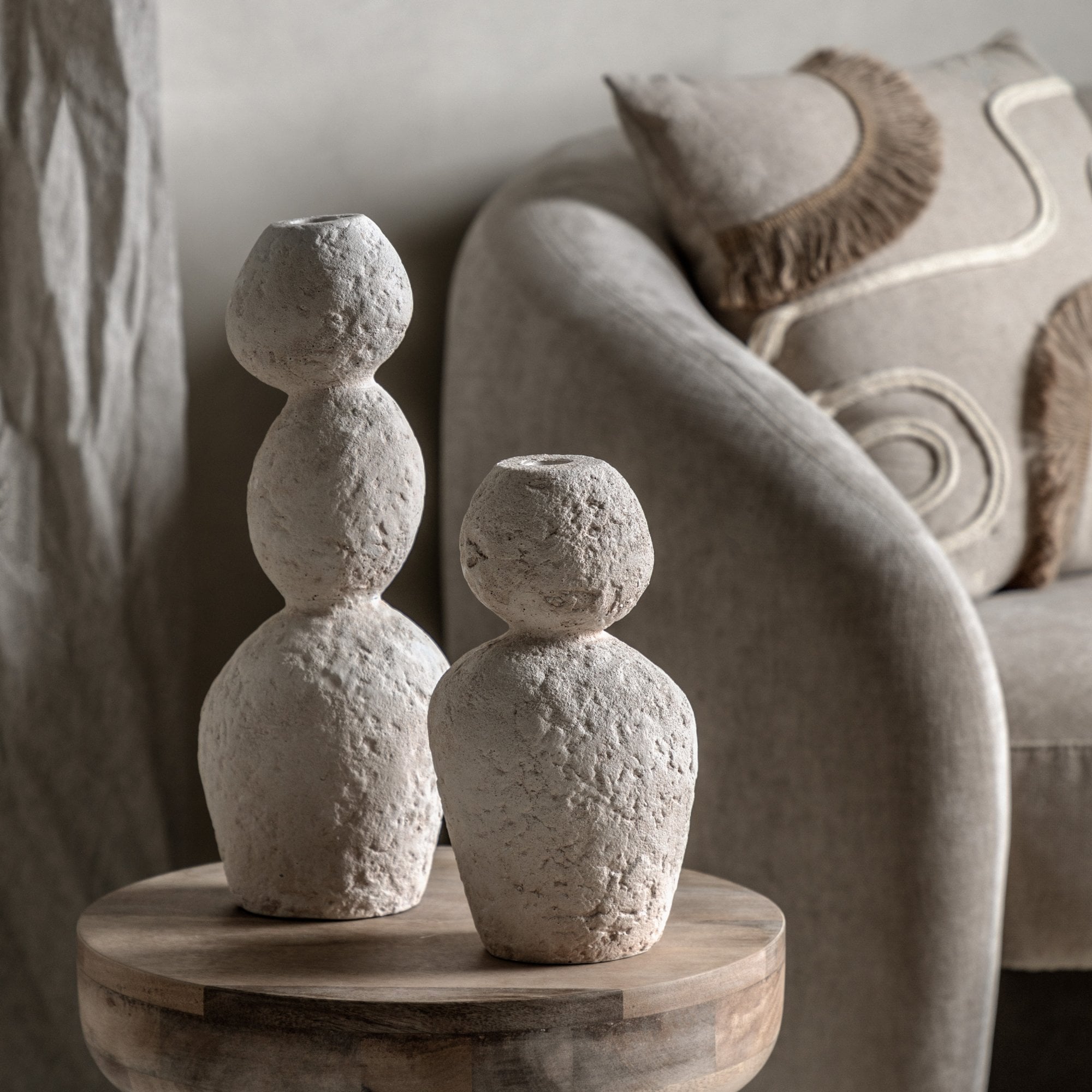 Two textured white stacked stone vases on a wooden side table next to an armchair.