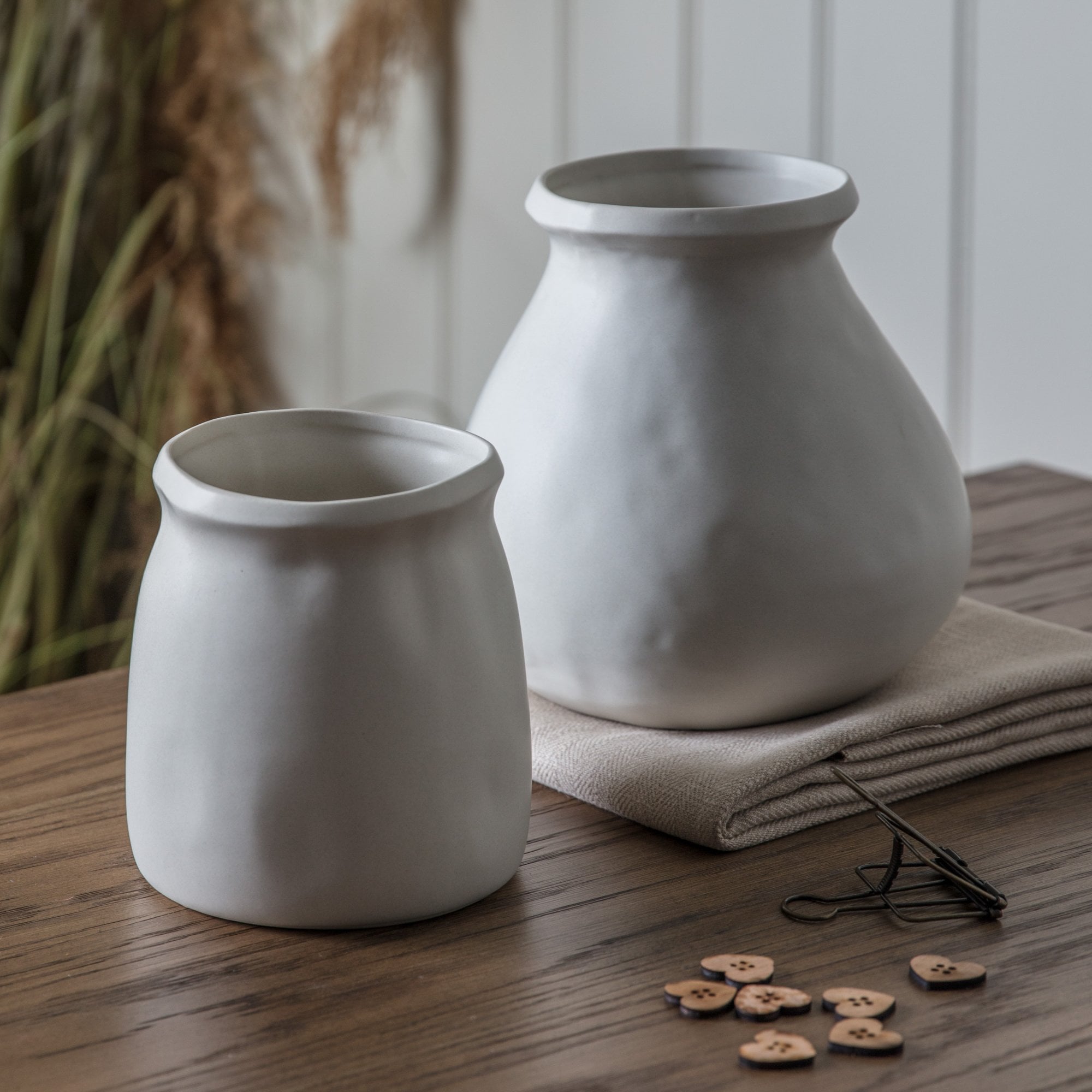 Two white vases of different sizes on a wooden table with dried grass in the background.