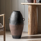 A tall, black and brown vase with two handles, placed next to a wooden console table.