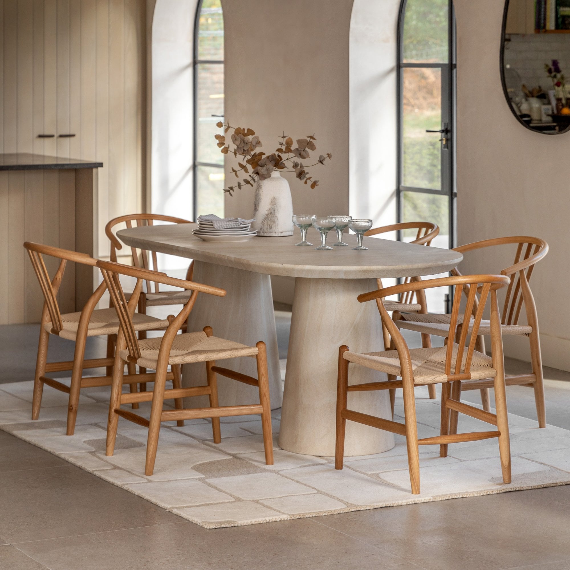 An oval dining table made of light beige stone surrounded by six wooden chairs with woven seats.