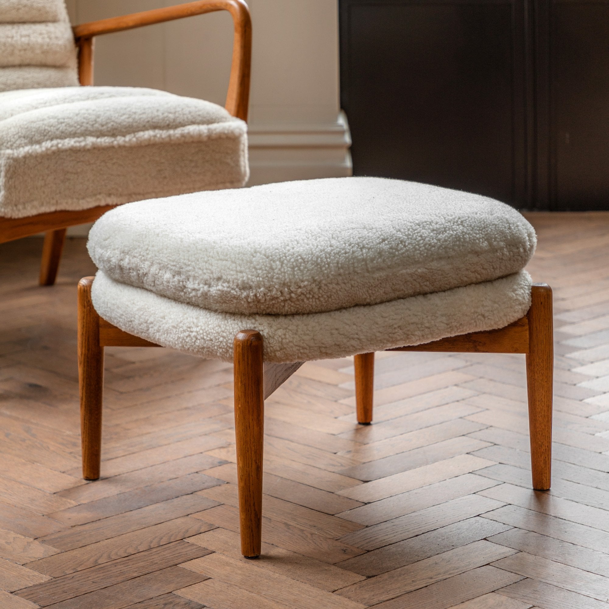 A white sheepskin footstool with a light wooden frame and legs on a herringbone floor.
