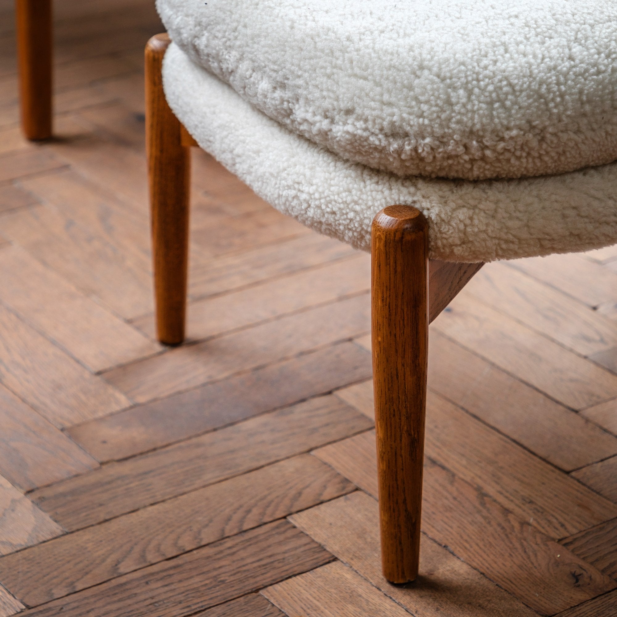 Close-up of the light wooden legs of a white sheepskin footstool on a herringbone floor.