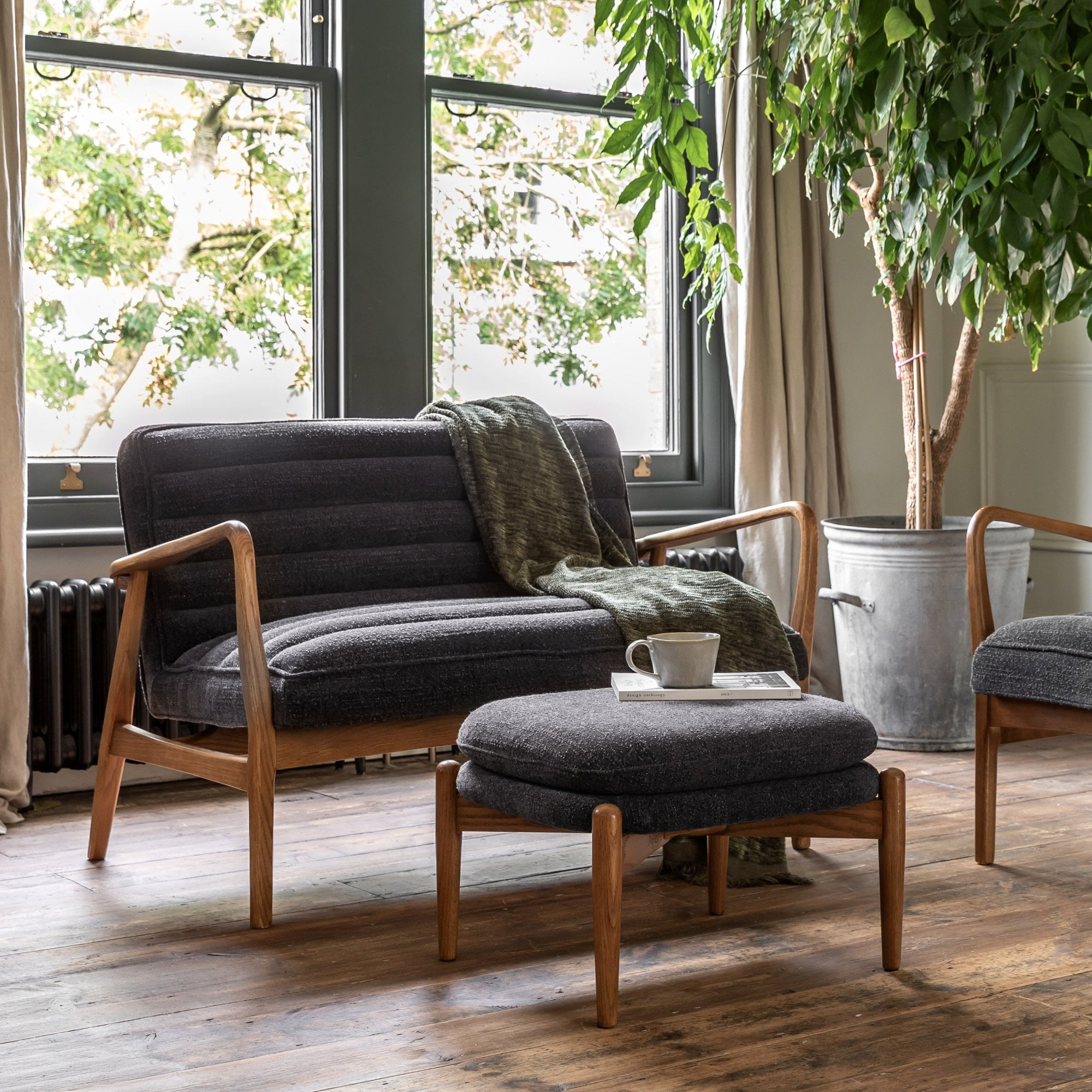 A mid-century modern grey upholstered ottoman in a living room with sofa and plant.