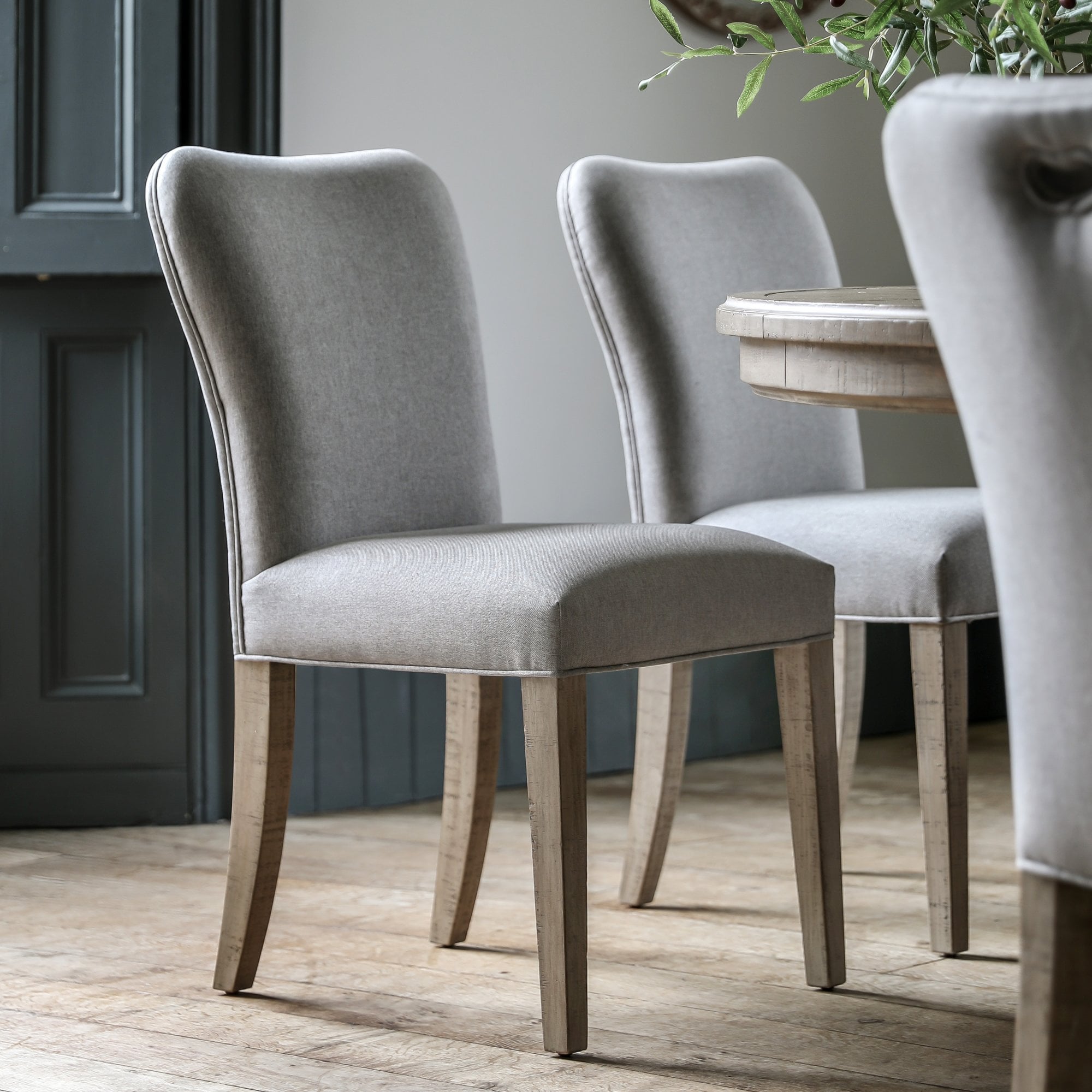 Four upholstered dining chairs with light wooden legs and a grey fabric finish arranged around a dining table.