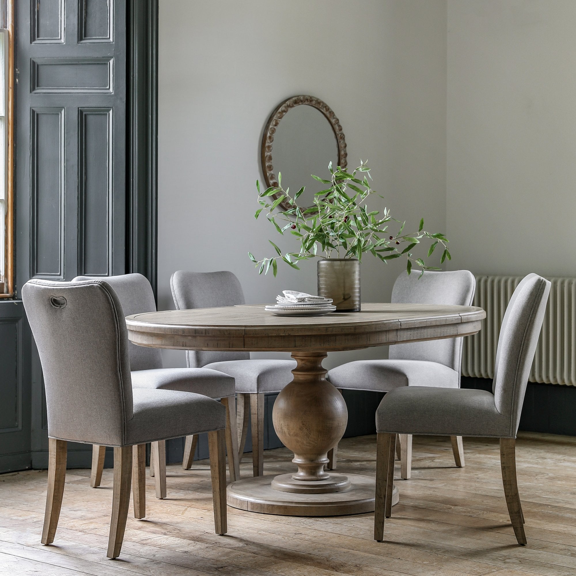 A round wooden pedestal dining table with four light grey upholstered chairs in a dining room.