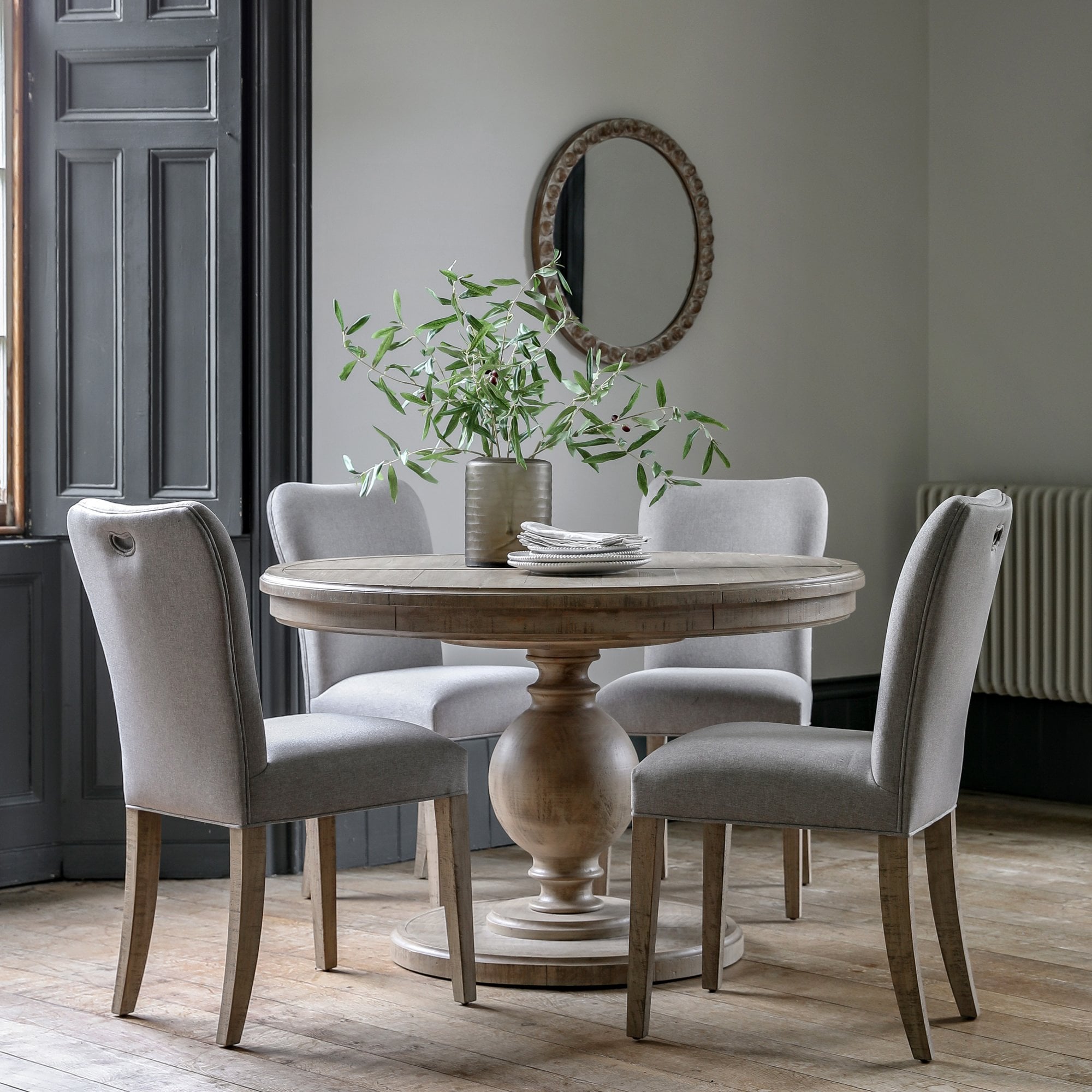 A round wooden pedestal dining table with four light grey upholstered chairs in a dining room.
