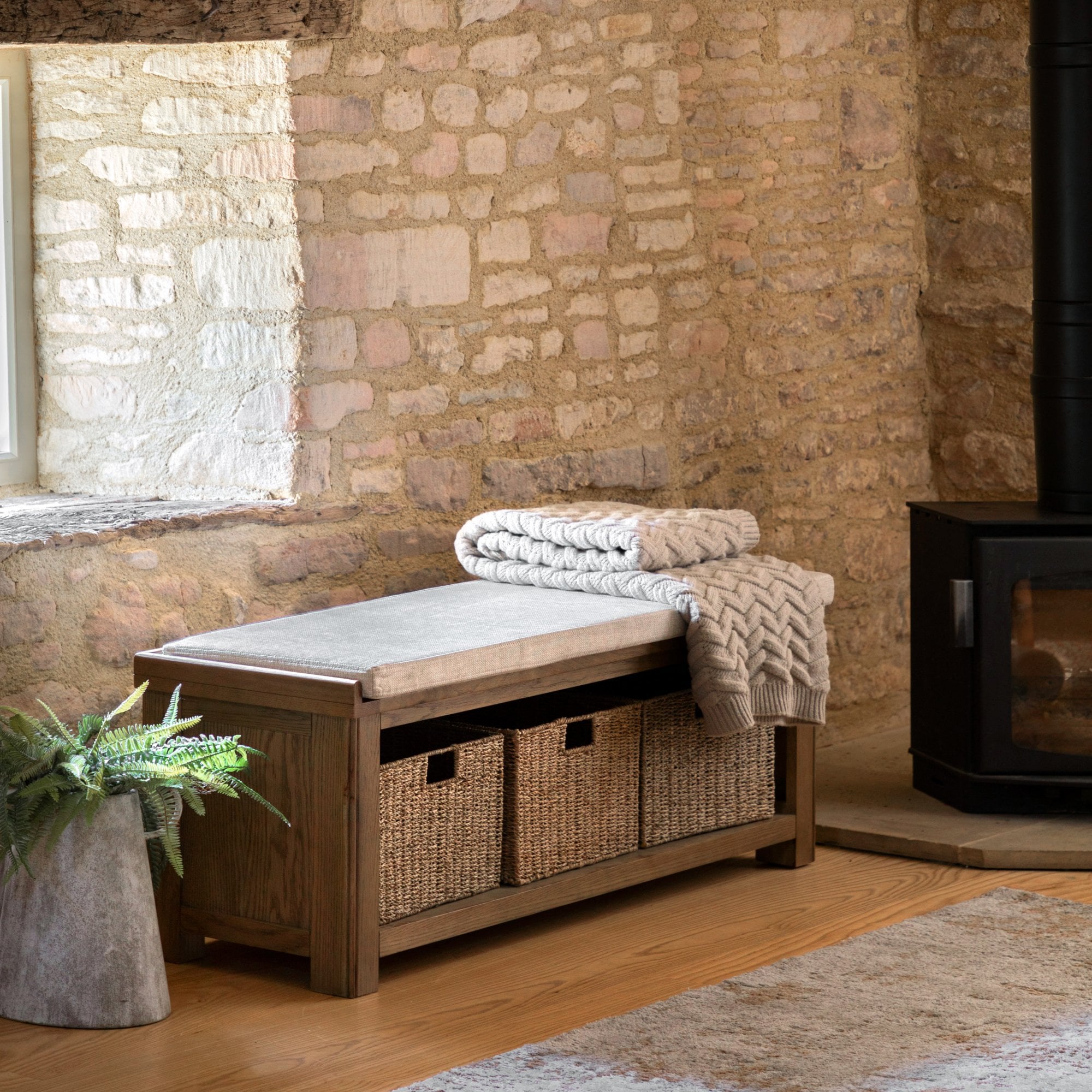 A wooden storage bench with a cushioned top, wicker baskets, and a throw blanket in a room.