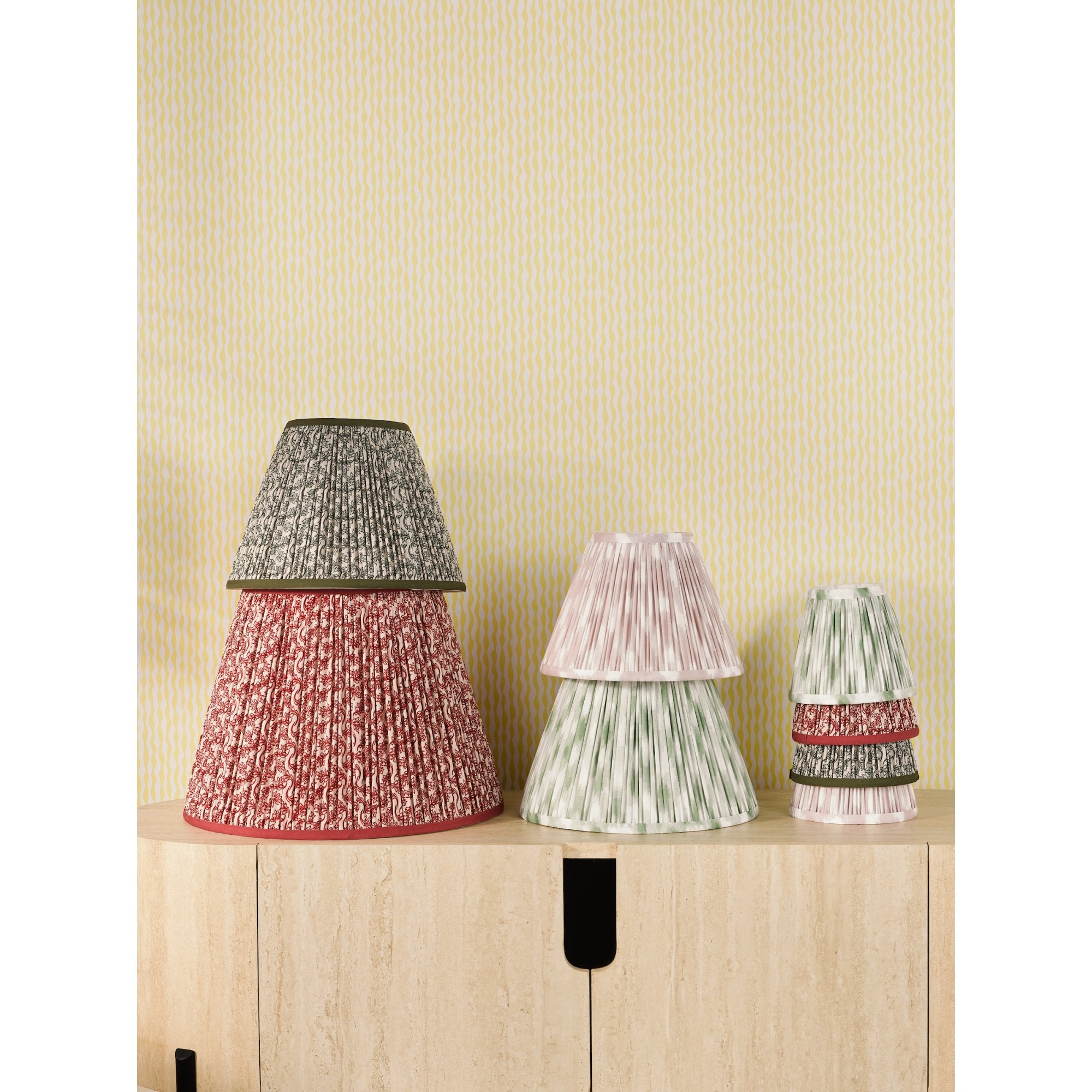 A light-yellow wallpapered room has various lamp shades in different sizes on a light-wood cabinet.