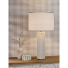 A white fluted table lamp on a wooden shelf, with a small vase and striped box beside it.
