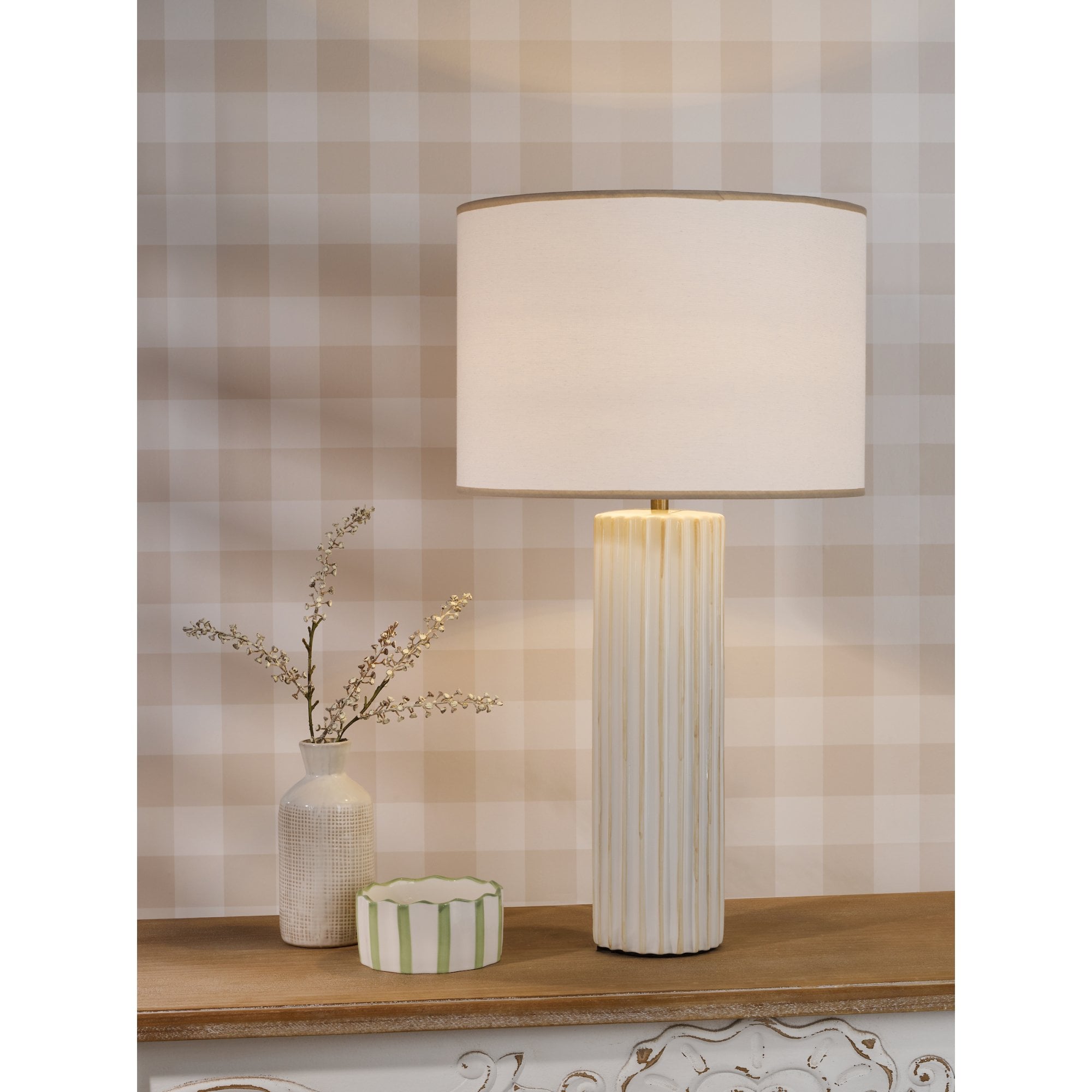 A white fluted table lamp on a wooden shelf, with a small vase and striped box beside it.