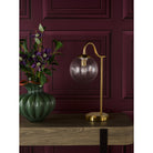 A table lamp with a brass finish and a clear glass shade next to a green vase.