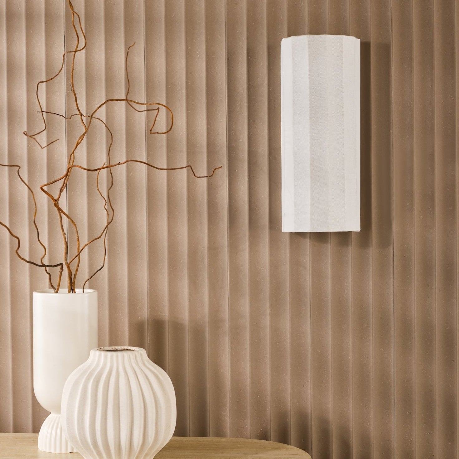 A white fluted wall light on a textured brown wall, with decorative vases on a table.