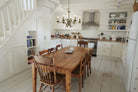 A brass chandelier hangs above a large wooden table in a white kitchen with stairs.