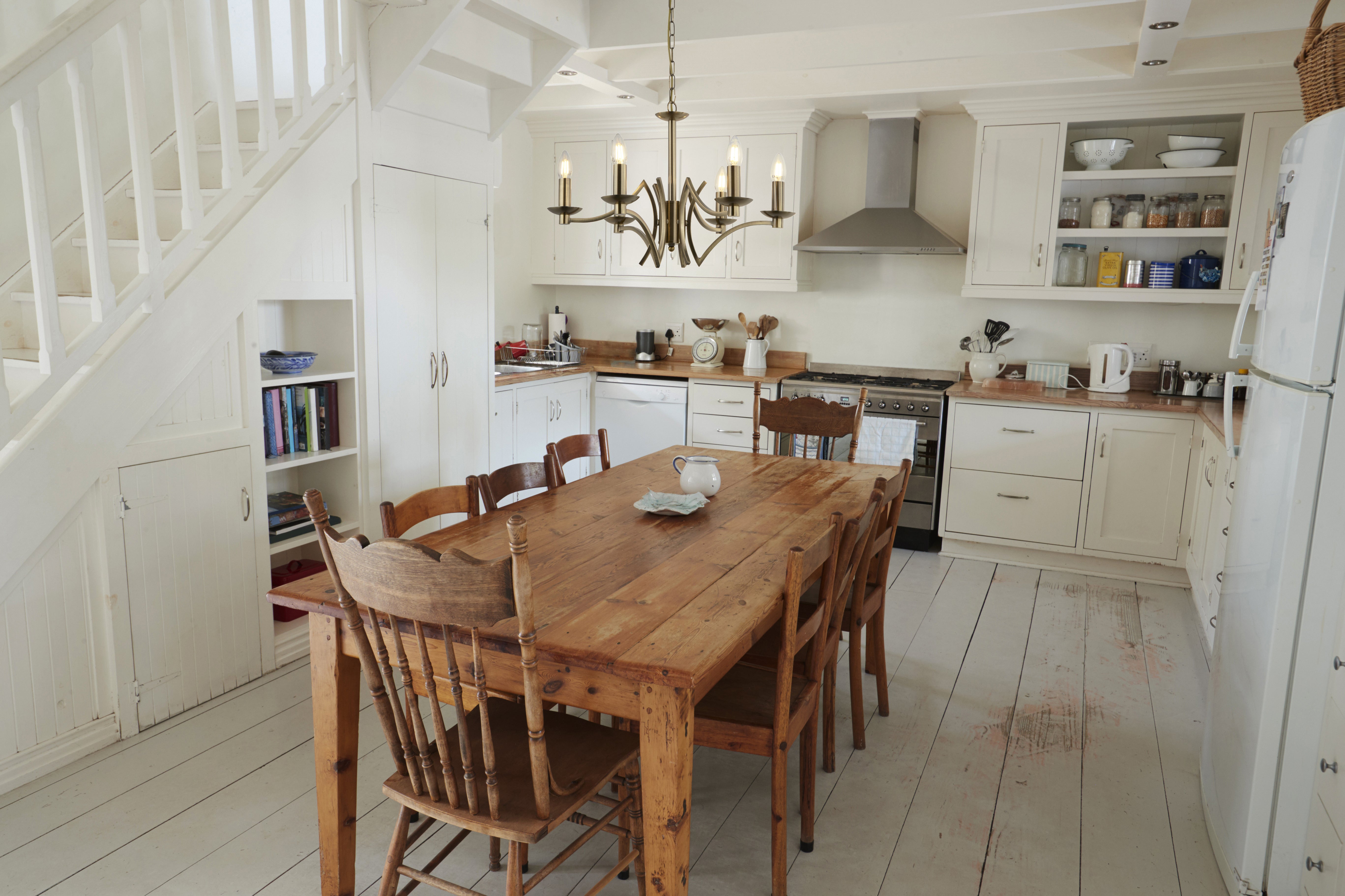 A brass chandelier hangs above a large wooden table in a white kitchen with stairs.
