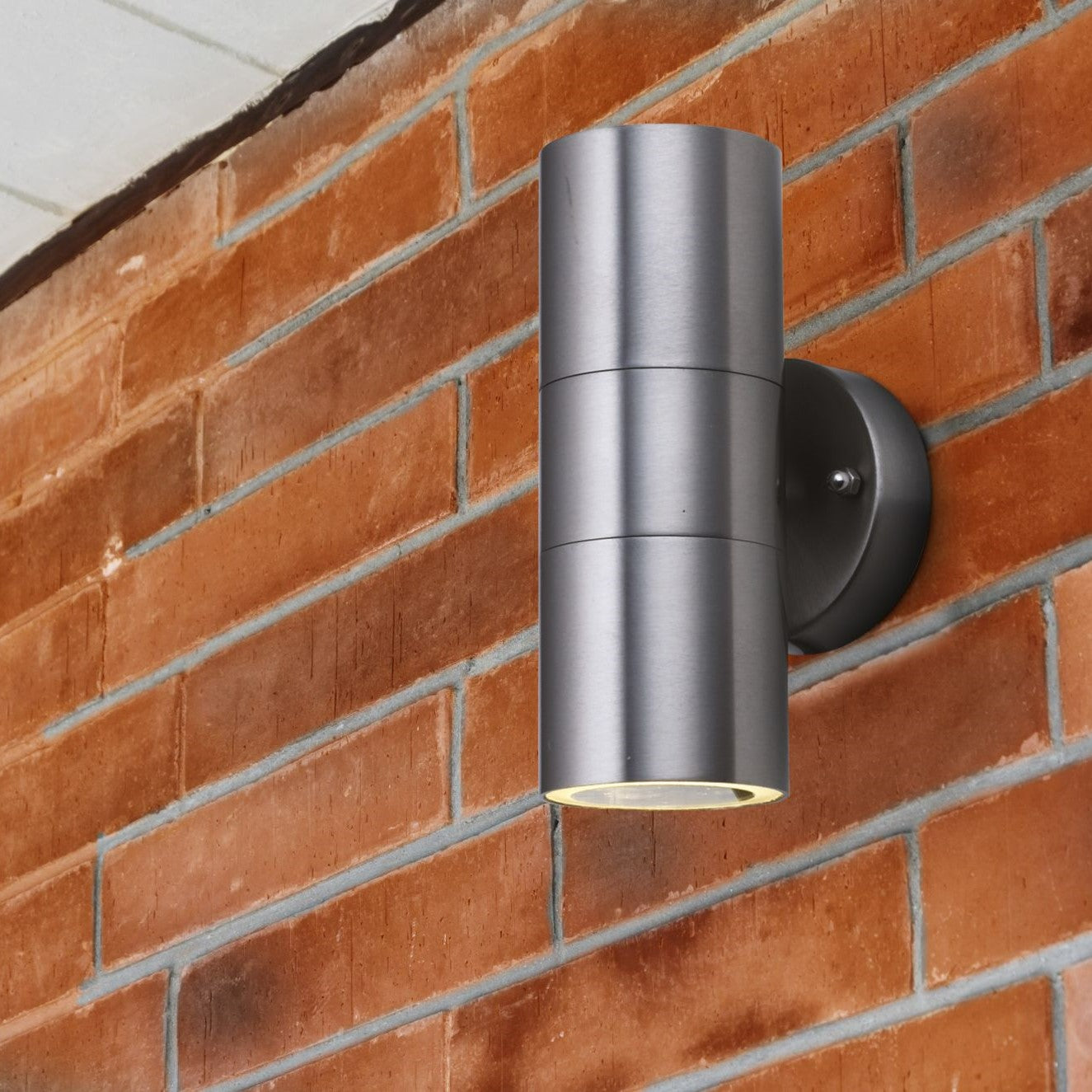 A modern, cylindrical brushed steel outdoor wall light installed on a brick wall.