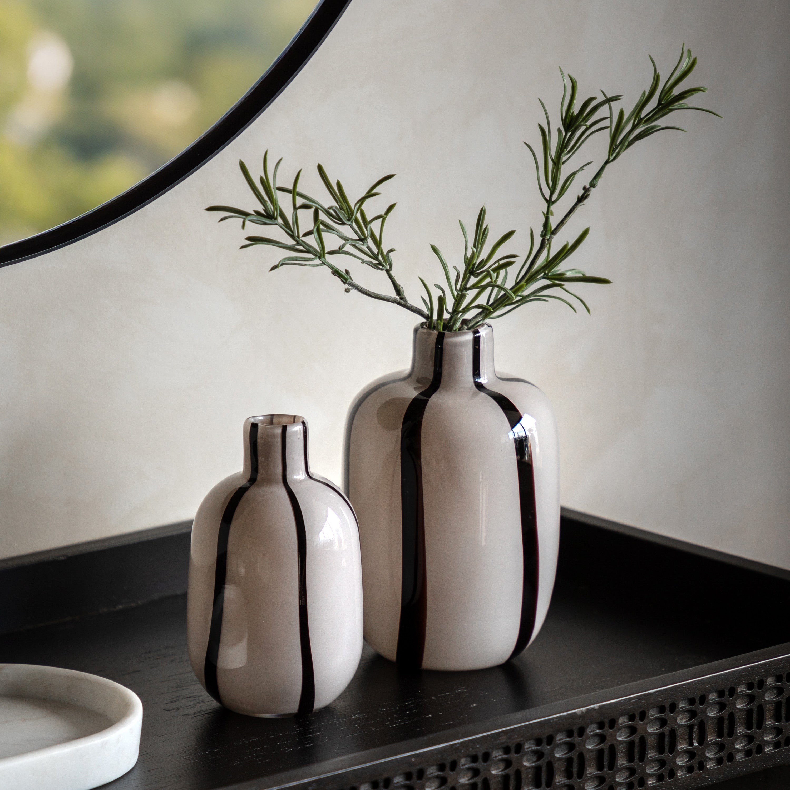 Two off-white vases with vertical black stripes, one larger with rosemary, on a dark table.