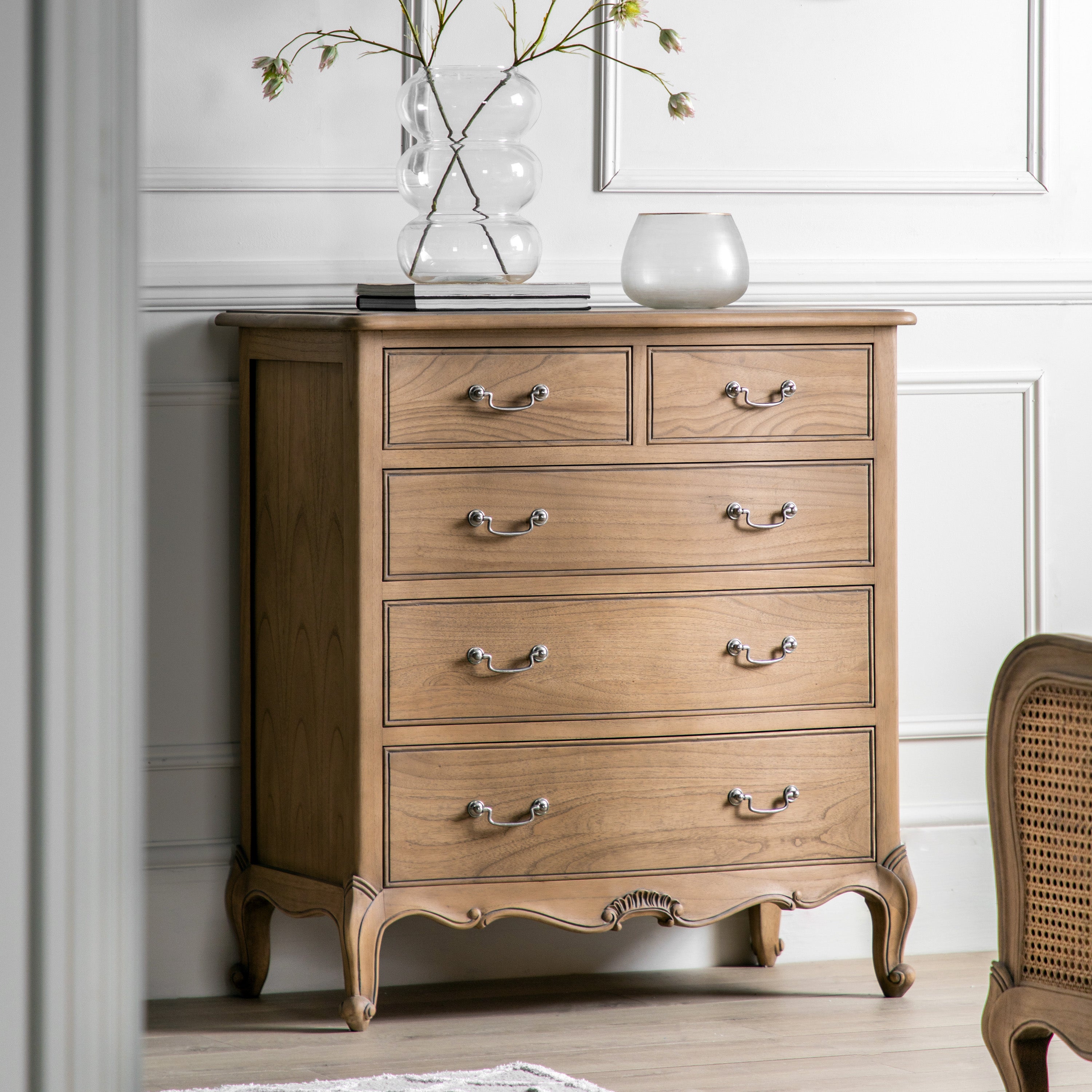 An ornate five-drawer chest of light wood with plants on top, in a room setting.