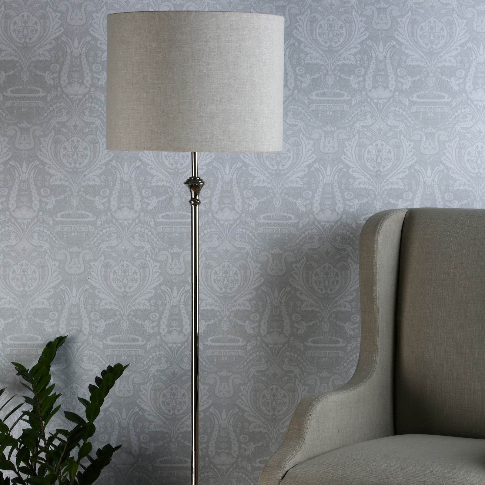 A floor lamp with a neutral shade and chrome stand, illuminating a patterned wall and a chair.