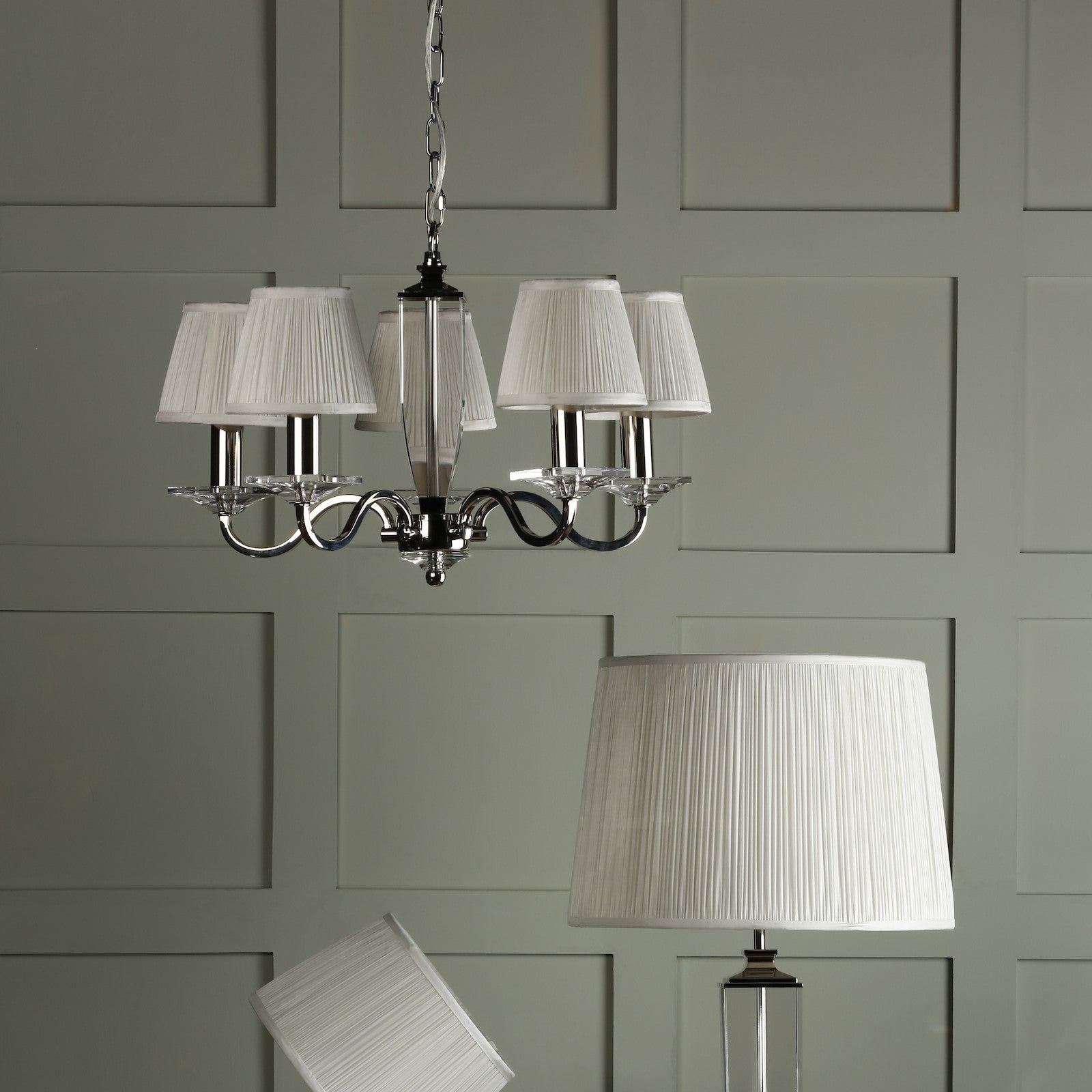 A chandelier with five pleated white lampshades hangs above a floor lamp with a matching shade.