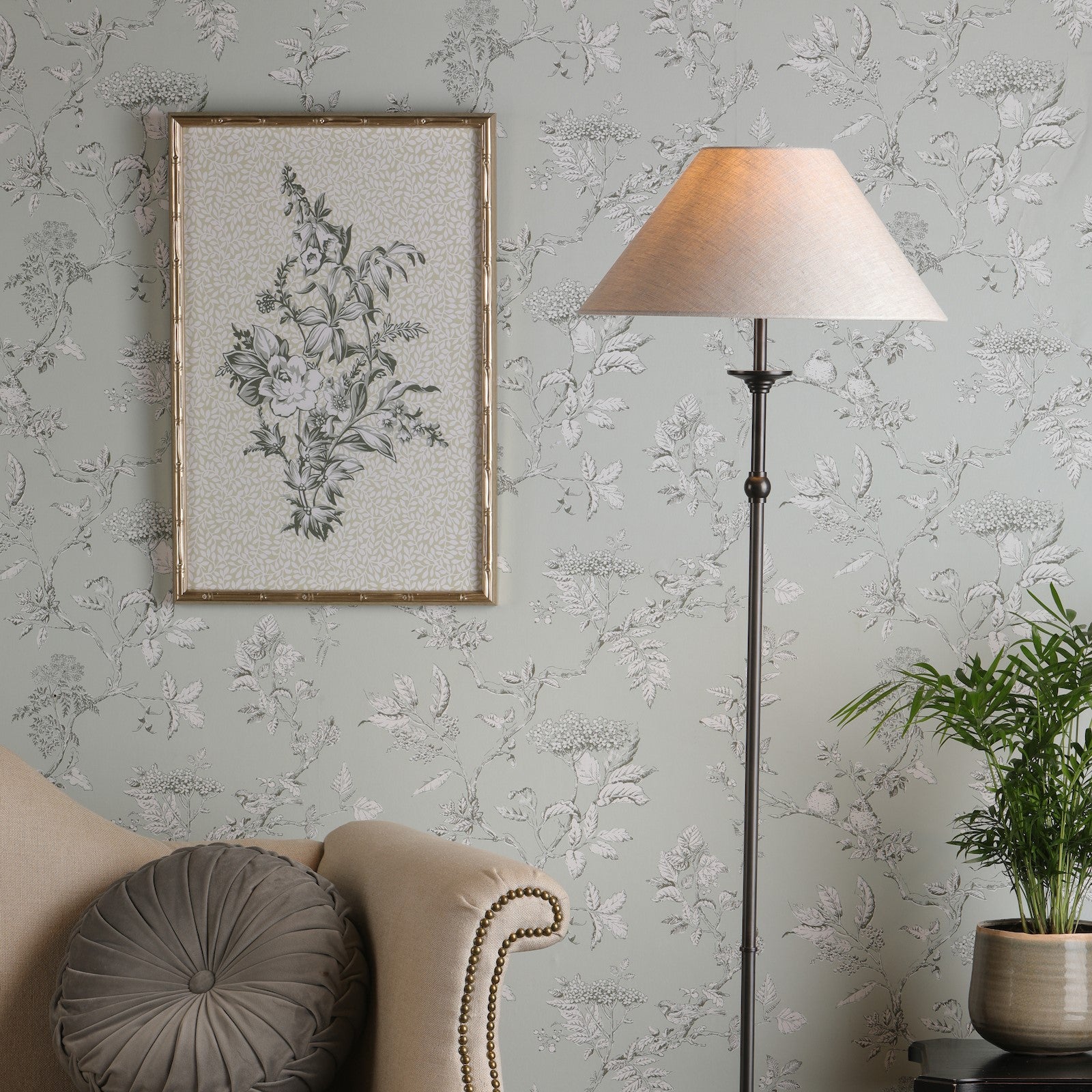 A floor lamp in a living room next to a beige sofa and patterned wallpaper. It illuminates.
