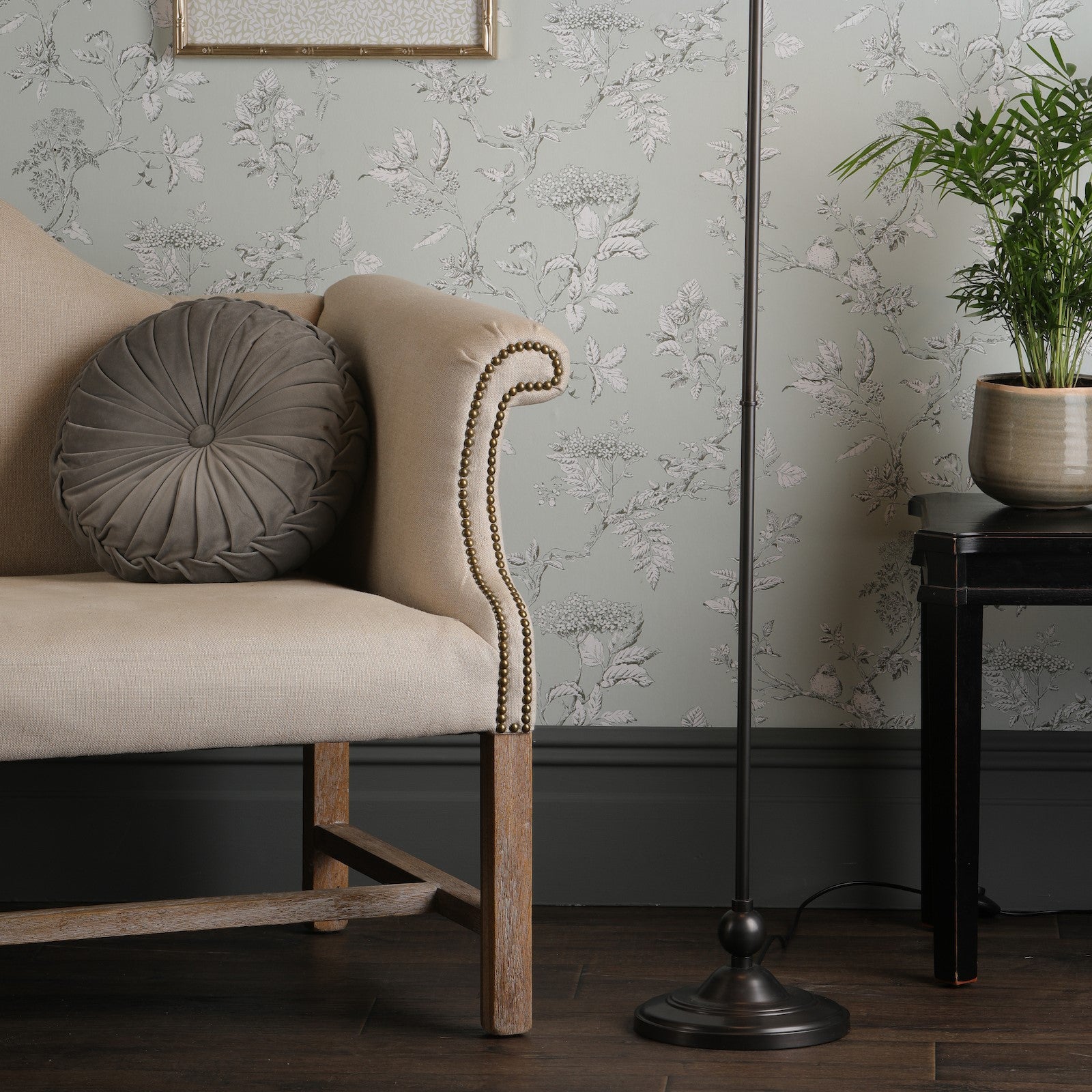 A floor lamp in a living room next to a beige sofa and patterned wallpaper. The light is off.