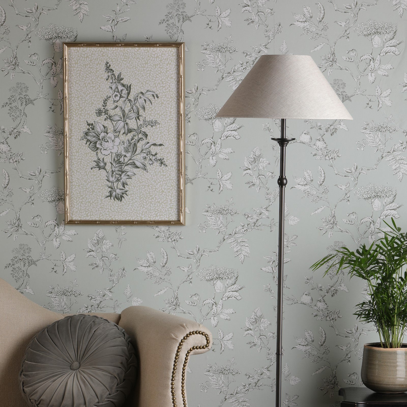 A floor lamp in a living room next to a beige sofa and patterned wallpaper. It illuminates.