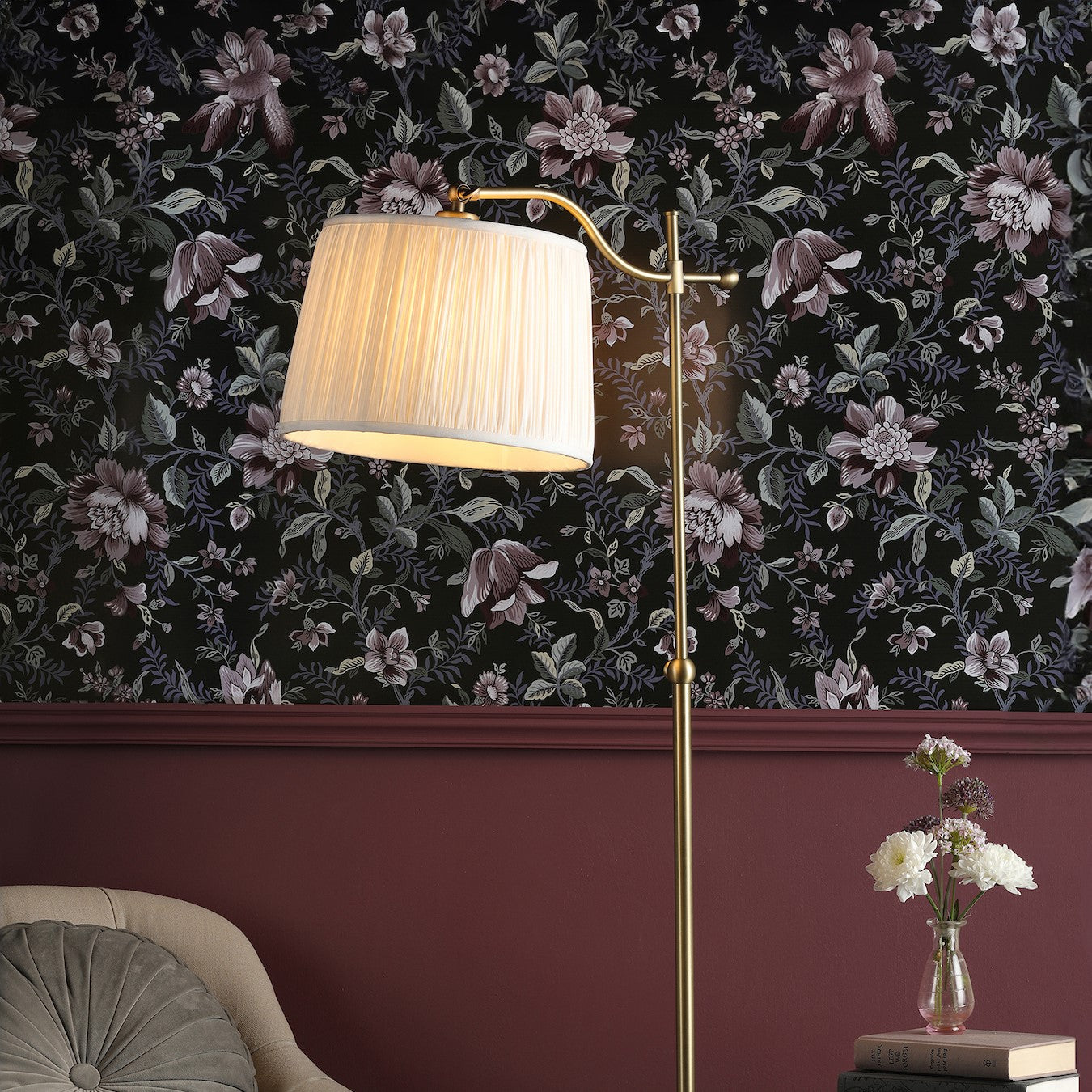 An elegant floor lamp with a pleated white shade illuminates a room with floral wallpaper.