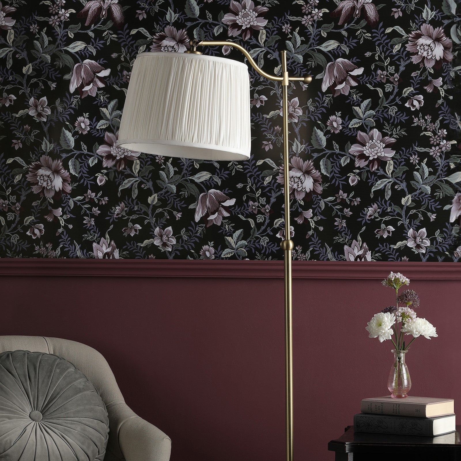 A brass-colored floor lamp with a pleated shade in a room with dark floral wallpaper.