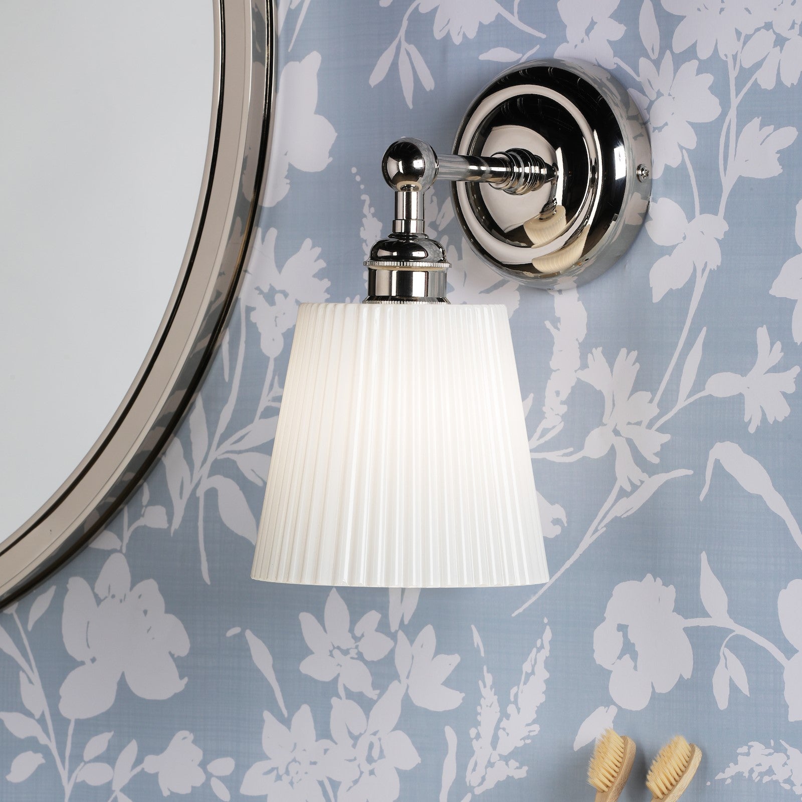 Polished nickel wall light with pleated shade illuminating a blue floral wallpapered bathroom.