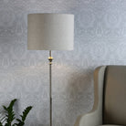 A floor lamp with a neutral shade and chrome stand, illuminating a patterned wall and a chair.