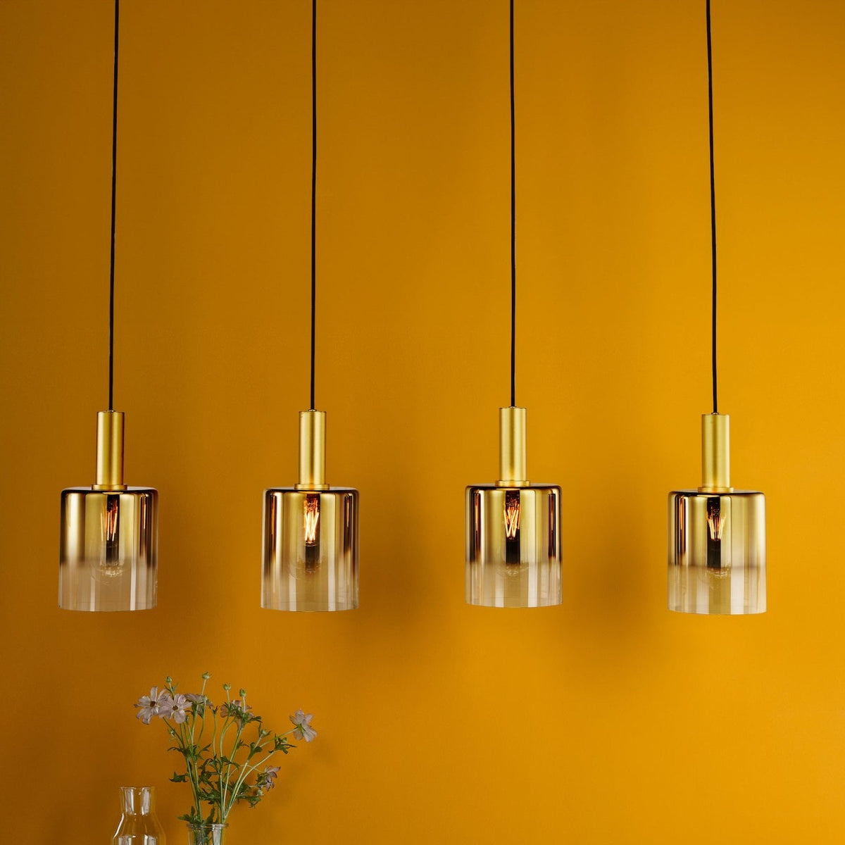 Four gold and clear glass pendant lights hanging over a wooden cabinet with a vase of flowers.