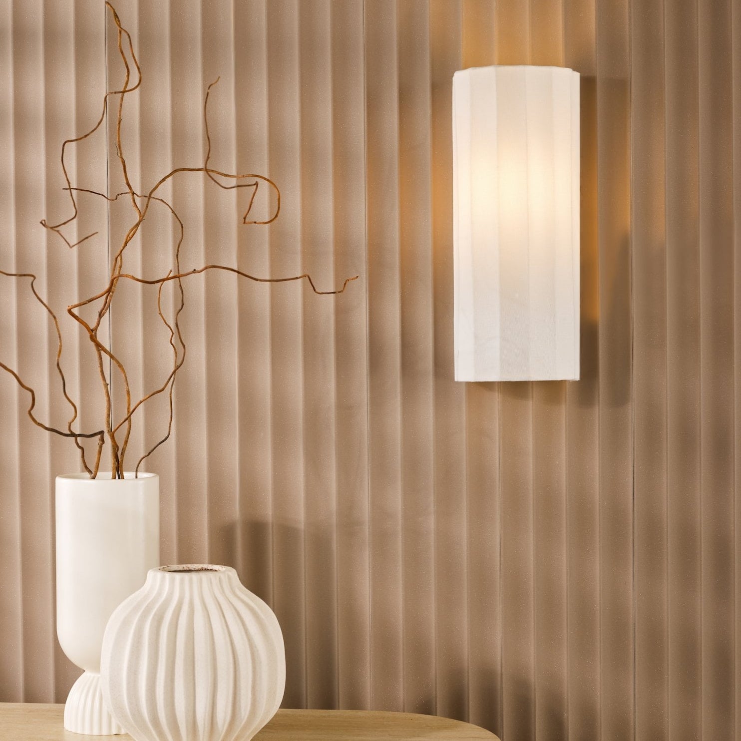A white fluted wall light illuminates a textured brown wall above a console table with vases.