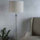 A floor lamp with a neutral shade and chrome stand, illuminating a patterned wall and a chair.
